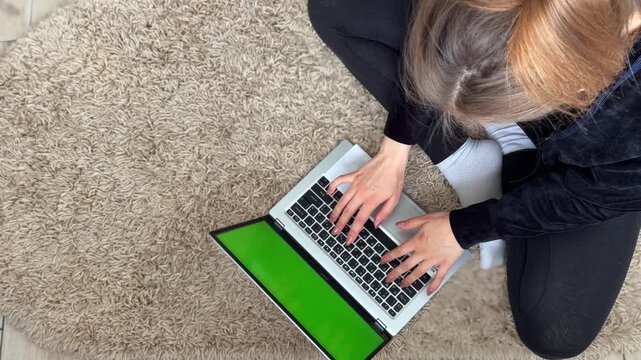 Freelancer woman typing on a laptop with a green screen, sitting on a round rug at home. Top down view of a student working remotely with a chroma key display notebook