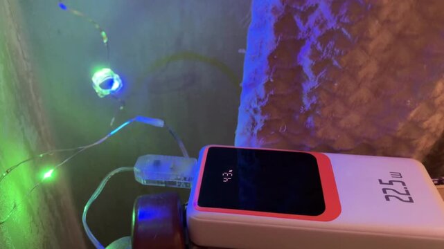 Portable power bank with digital display charging a colorful flashing led light strip, showcasing the device's battery level and modern electronic technology concept