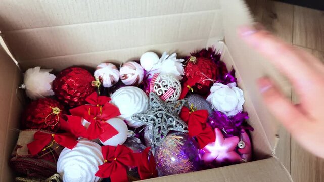 Woman hands opening cardboard box full of various red, white, and purple christmas tree decorations and ornaments, preparing for winter holidays and festive celebrations