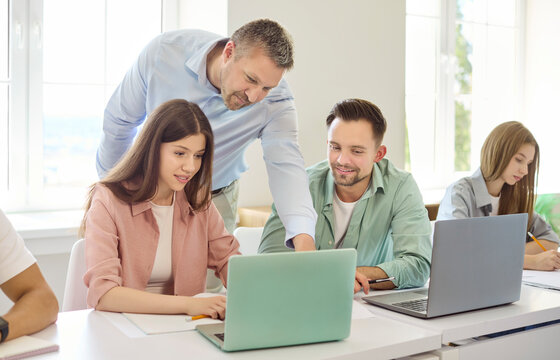 Students and teacher in classroom with laptop. College peers get guidance as the teacher points at screen, aiding teamwork and problem solving. Clear concept of mentoring teamwork and learning.