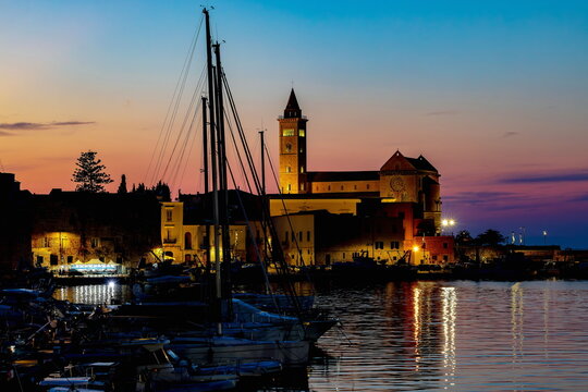 Contro luce del fronte mare della  cattedrale di Trani ( Puglia , Italia ) al tramonto con sagome degli alberi di barca a vela 