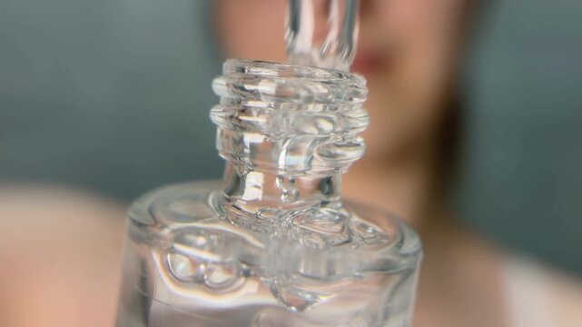 Close up of a beauty cosmetic serum dropper taking hyaluronic acid from a glass bottle, showing the transparent liquid with bubbles and a woman in the background
