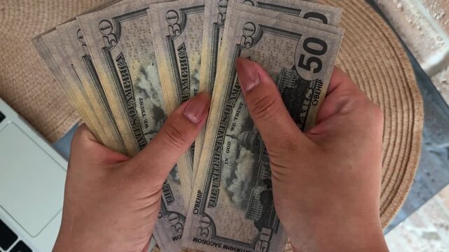 Woman's hands counting fifty dollar bills, top view of a freelancer calculating salary or savings, financial success and personal finance management at a desk with a laptop