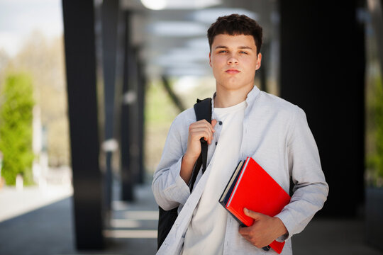 Young man stands outside a building with books and a backpack during daytime in a modern area