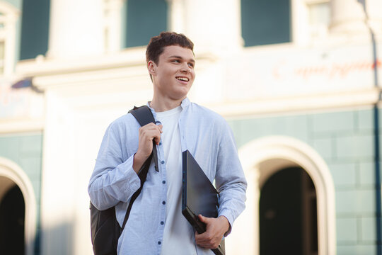 Student walks outside of a building while carrying a laptop and smiling in a city setting during the day