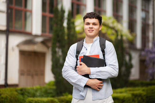 Student holds books outside a school during a sunny day while smiling at the camera