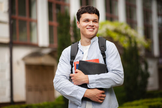 Young man stands outside holding a notebook and smiling in front of a school building during a sunny day