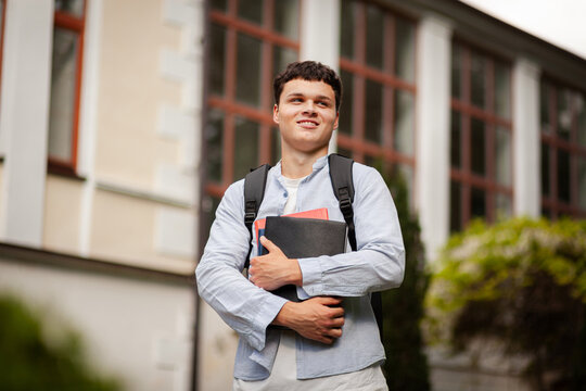 Student stands outside school holding books and smiles during mid-morning break with trees nearby
