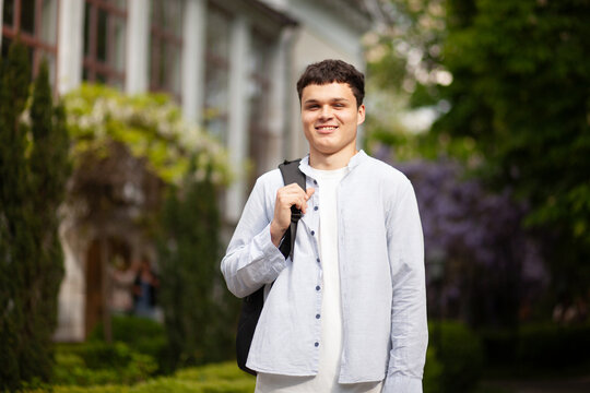 Smiling young man stands outside building with greenery and purple flowers on a clear day in a park setting