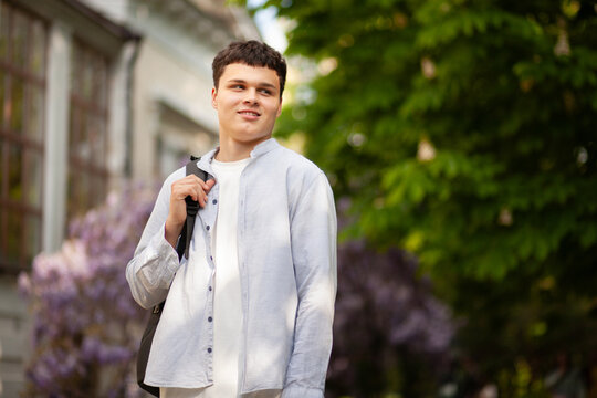 Young man walks in a park during spring with a bag over his shoulder and smiles at the camera on a sunny day