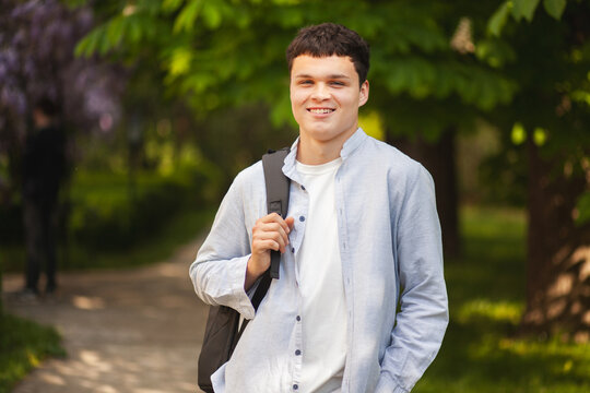 Young man stands on a path in a green park with trees and flowers while smiling in daytime