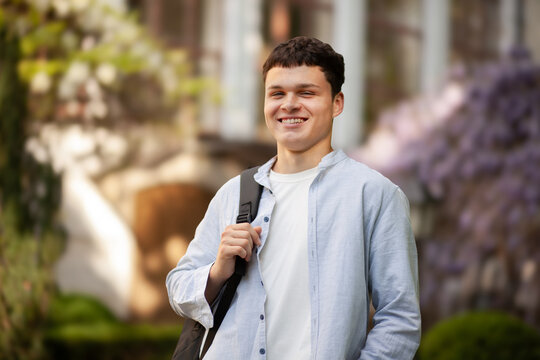 Young man smiles while standing outside in a garden during daytime with blooming flowers around him