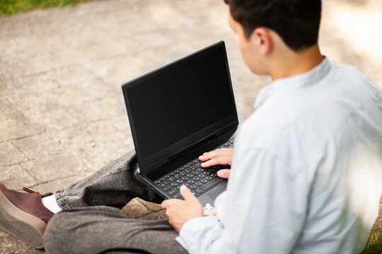 Young person works on laptop while sitting on ground in outdoor space during daytime with natural light around them