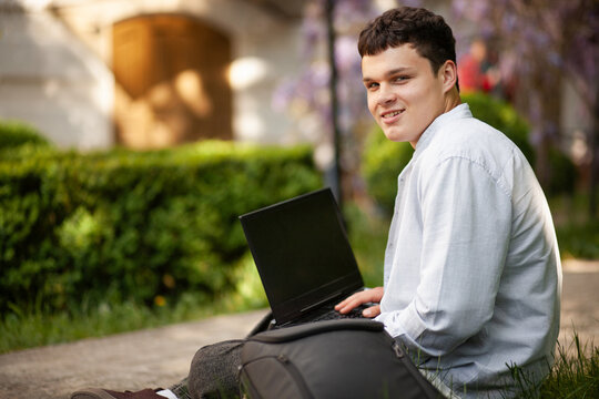 Young person works on laptop outside near bushes and flowers in daylight with a slight smile on their face and a backpack by their side