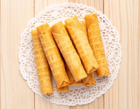 Golden crispy rolled egg wafer cookies stacked on white paper doily on light wooden table. Traditional homemade dessert, top view.
