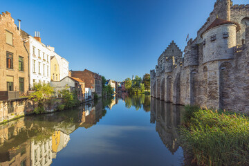 Fototapeta premium Gravensteen, a medieval castle in the city of Ghent in Belgium, Europe. Historic European Canal with Clear Blue Sky, Stone Buildings, and Serene Water Reflections on a Sunny Day