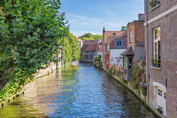 Obraz premium Bruges town in Belgium, Europe. Charming canal scene with historic buildings and lush green trees along the tranquil waterway under a bright blue sky on a sunny day