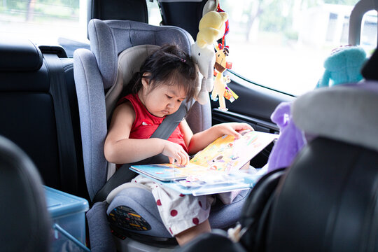 Cute Asian Little Girl Reading a Book in a Car Seat During Travel,A cute Asian little girl sitting safely in her car seat in the backseat of a car, focusing on reading a colorful storybook while trave