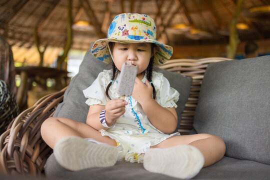 Cute Little Girl Eating Ice Cream on a Sofa,A cute little Asian girl wearing a patterned bucket hat sitting comfortably on a rattan sofa and enjoying a delicious ice cream pop.