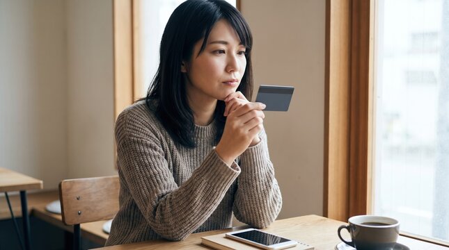カフェの窓際でクレジットカードを手に外を眺める思案顔の日本人女性、スマートフォンを置いたテーブル、カード利用を考えるイメージ
