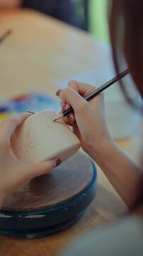 Close-up of female artist&rsquo;s hands sketching design with pencil on handmade ceramic pottery before painting &ndash; creative hobby, traditional craft and artistic expression in calm studio workshop setting
