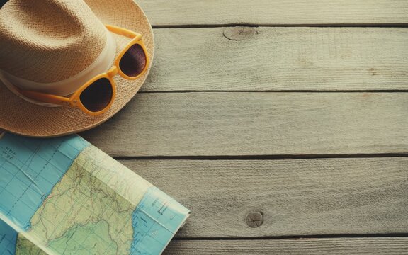 Travel essentials including a straw hat, orange sunglasses, and a folded paper map placed on a rustic wooden table, ready for summer vacation and holiday planning