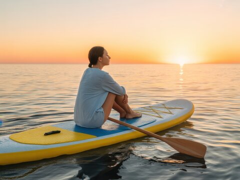 Young woman sitting on a stand up paddle board in the middle of a calm ocean during a golden sunset while enjoying a peaceful moment of reflection