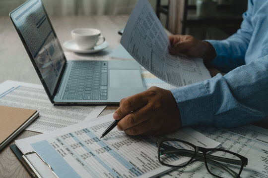 Business person reviews printed spreadsheets beside a laptop displaying charts, checking figures and comparing documents for data analysis, dashboard reporting, and business data management.