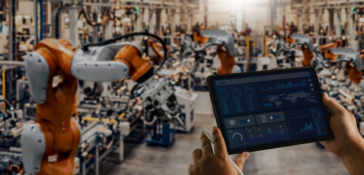 Hands holding a tablet monitor robotic arms in a smart factory, using a digital dashboard for automated manufacturing control, production analysis, and industrial operations management.