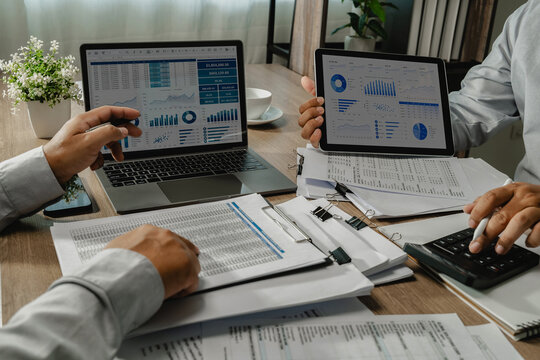 teamwork review printed reports, charts on a laptop and tablet, and calculator figures at a desk, comparing financial data for report analysis, dashboard reporting, and finance review.