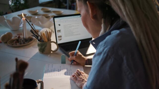 Woman potter plans tasks in notepad near laptop among pottery