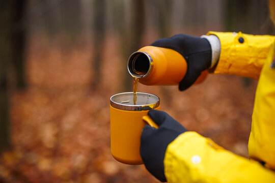 Hiker pouring hot drink from thermos into travel mug in autumn forest. Refreshment during hiking outdoors