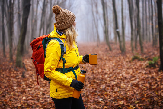 Woman hiker in yellow jacket with backpack holding thermos and travel mug in autumn forest. Refreshment during trekking in cold foggy weather