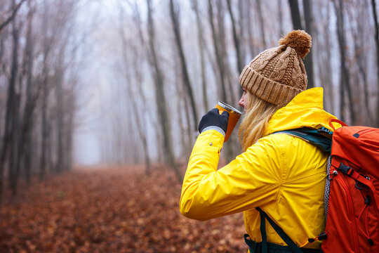 Woman hiker in yellow jacket drinking hot tea from travel mug at forest. Refreshment during autumn hiking in cold misty weather