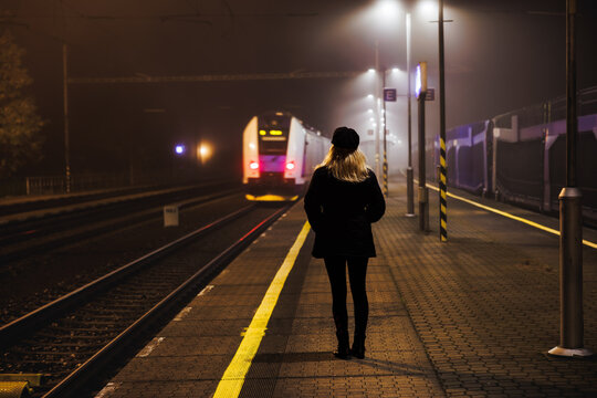Woman looking at leaving train at empty railway station at night. City life and commute by public transportation