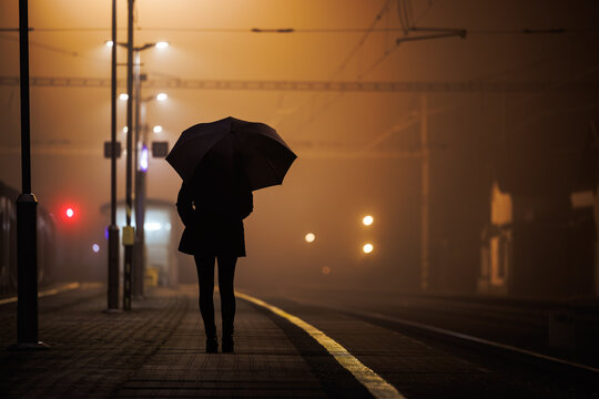 Railroad station at night. Dark silhouette of woman with umbrella waiting for train on empty railway station platform