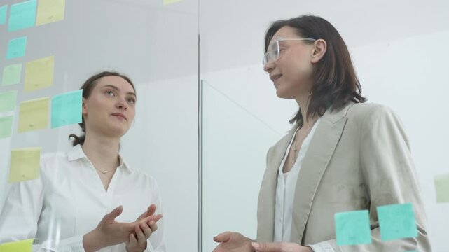 Diverse businesswomen collaborating and brainstorming innovative ideas while sticking and arranging colorful post it notes on a clear glass wall in a modern office meeting or workshop