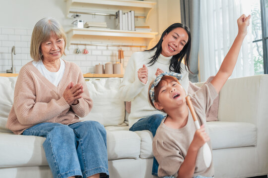 A little girl sings into a wooden spoon microphone while her mother dances and grandmother claps along on the sofa a playful, joyful family moment of impromptu music and shared laughter at home.