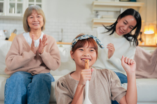 A little girl sings into a wooden spoon microphone while her mother dances and grandmother claps along on the sofa a playful, joyful family moment of impromptu music and shared laughter at home.
