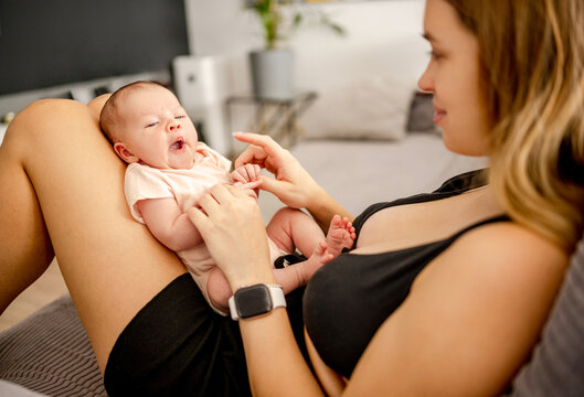Young mother lovingly touches newborn infant baby while sitting on bed at home during afternoon