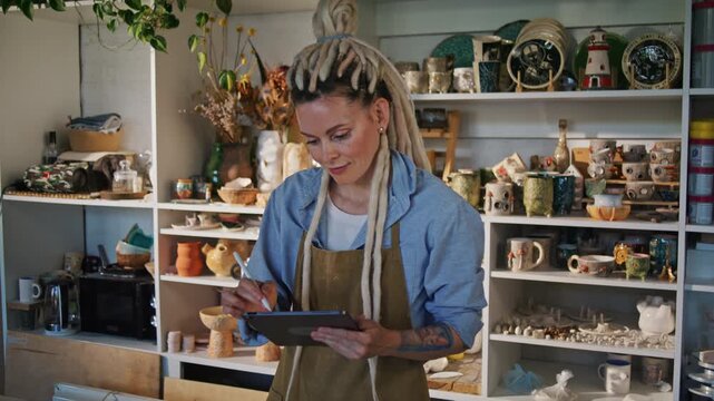 Lady potter writes workshop notes on tablet among ceramic shelves