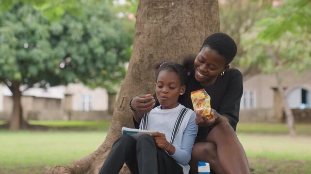 Black mother sharing snack and tablet, quiet picnic under tree with daughter focused on screen, gentle feeding gesture and warm tutoring moment, water bottle at roots and soft afternoon light