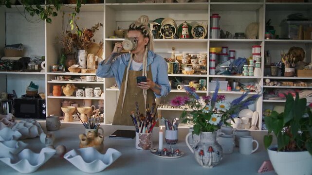 Lady potter drinks from handmade mug while checking smartphone
