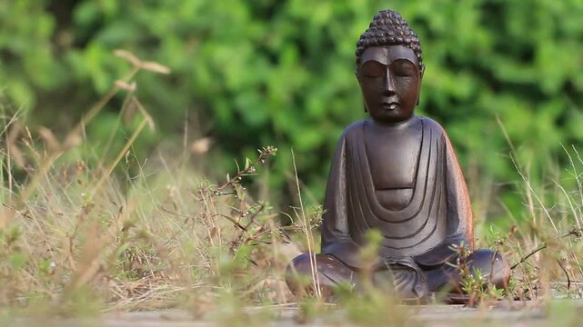 Small wooden buddha statue sitting in peaceful meditation pose among wild dry grass with green background.