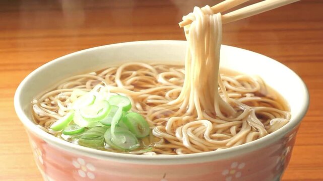 Chopsticks lifting steaming hot soba noodles from floral ceramic bowl on wooden table in cinematic close up.