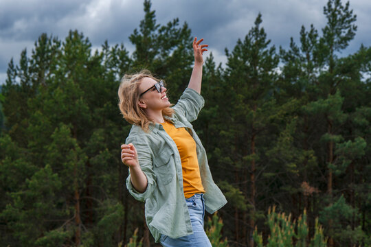 Happy woman in sunglasses standing with raised arm against green pine forest. Cheerful outdoor recreation and active summer lifestyle concept