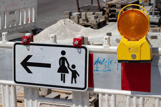 Traffic sign indicating left turn for pedestrians with child, illuminated warning light mounted on construction barrier in urban setting with building materials nearby