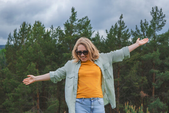 Cheerful woman in sunglasses standing with wide open arms in green forest. Travel freedom inspiration and positive summer emotion concept