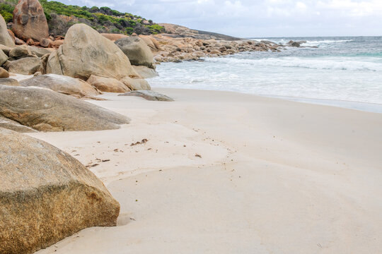Coastal headland with distinctive red gneiss rocks beside a sandy beach under natural daylight. A rugged shoreline scene showing rock formations, ocean waves, and coastal landscape textures.