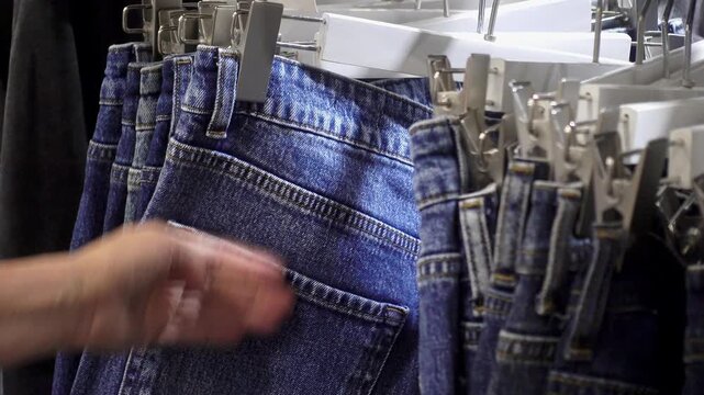A shopper's hand examines blue denim jeans hanging on clips and hangers in a retail clothing store display, showing casual apparel selection and shopping activity